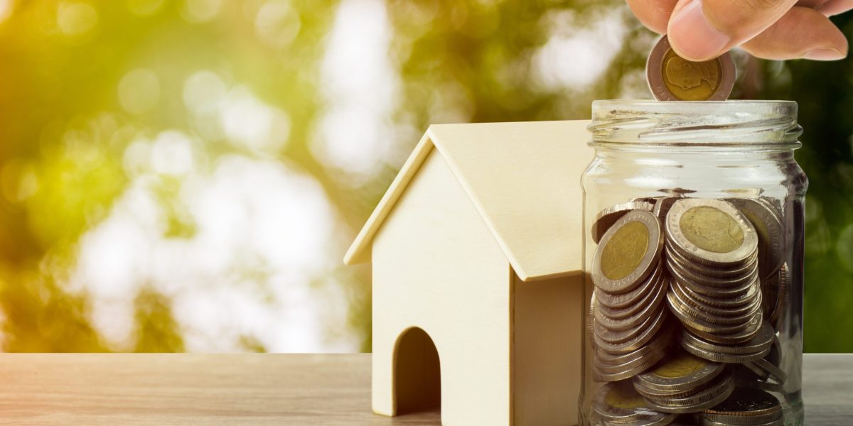 Property investment or saving money for buy home. A businessman hand holding coin over glass jar and resident house model on wooden table.